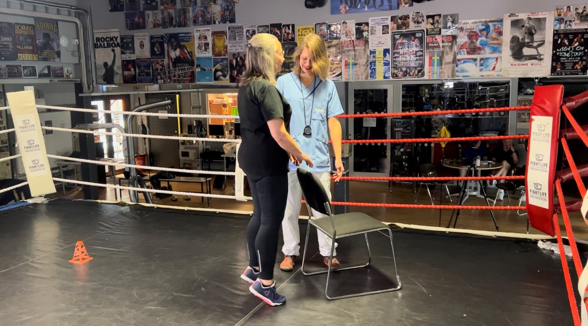 En äldre kvinna står i en boxningsring tillsammans med en fysioterapeut. Den äldre kvinnan gör tåhävningar. 