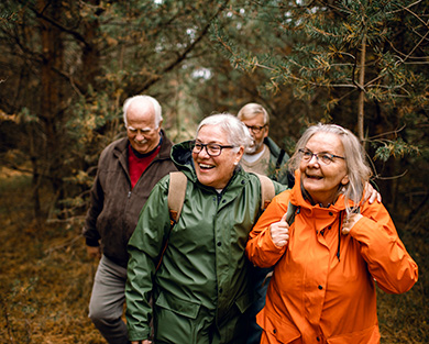 Fyra seniorer är ute i en skog och promenerar. Det tycks vara höst för löven är orangea. Personerna ler och är glada. 